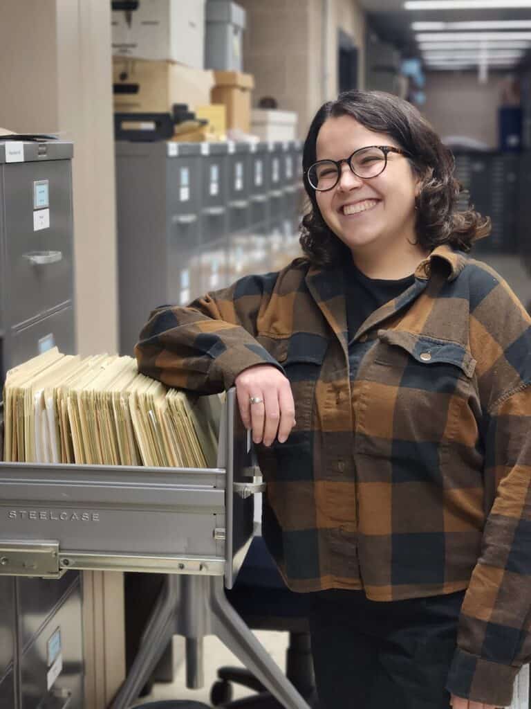 Young woman wearing a flannel shirt, black rimmed glasses, and short brown hair smiles to the camera. One arm leans on an open filing cabinet drawer. 
